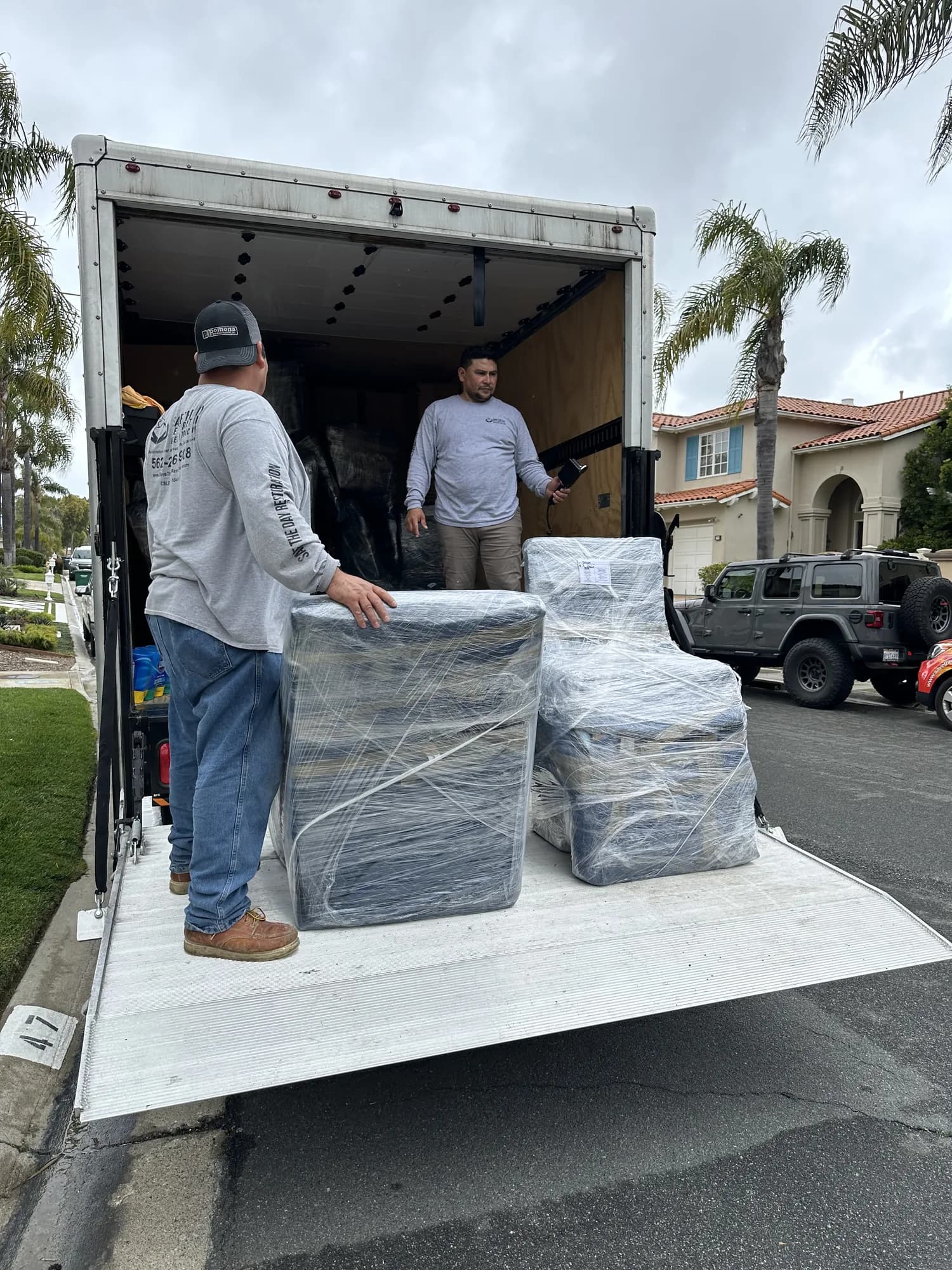Two crew members loading shrink-wrapped furniture onto a box truck lift gate during a pack-out